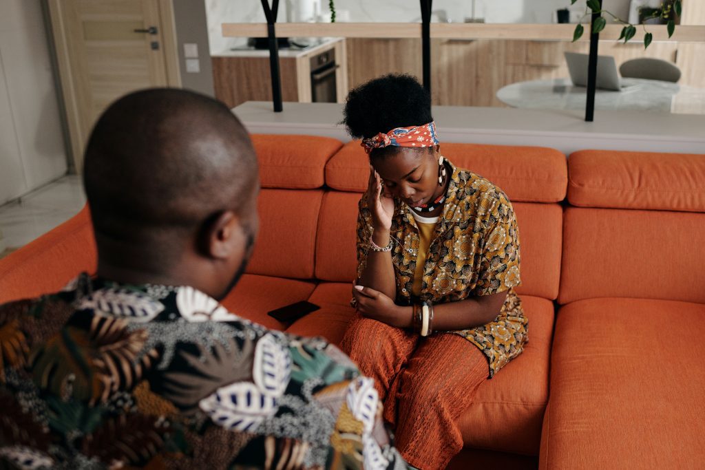 Lady Sitting On Red Couch With Her Head In Her Hands Talking To Her Therapist, A Man Facing Her In A Colourful Shirt